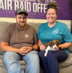 Man and woman seated with a puppy. Man wears hat and brown shirt, woman in teal shirt. All are smiling.