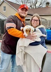Two people holding a dog wrapped in a blanket outside. The man wears a brown and orange jersey.