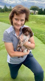 Woman kneeling, holding a small brown and white puppy on green grass.