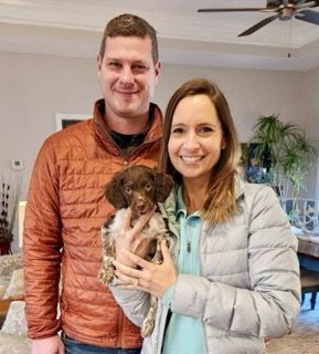 Couple indoors holding a small brown and white dog; man in orange jacket, woman in silver jacket.