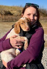 Woman holding a cream and brown puppy outdoors. Both smiling in front of a rocky hillside.