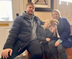 Family seated with puppy indoors. Man and woman in winter coats, child smiles, holding the dog.