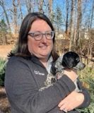 Woman with glasses smiling and holding a small black and white dog outdoors.