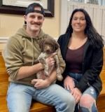 A man in a green hoodie holding a small dog, and a woman in a black jacket sitting together.