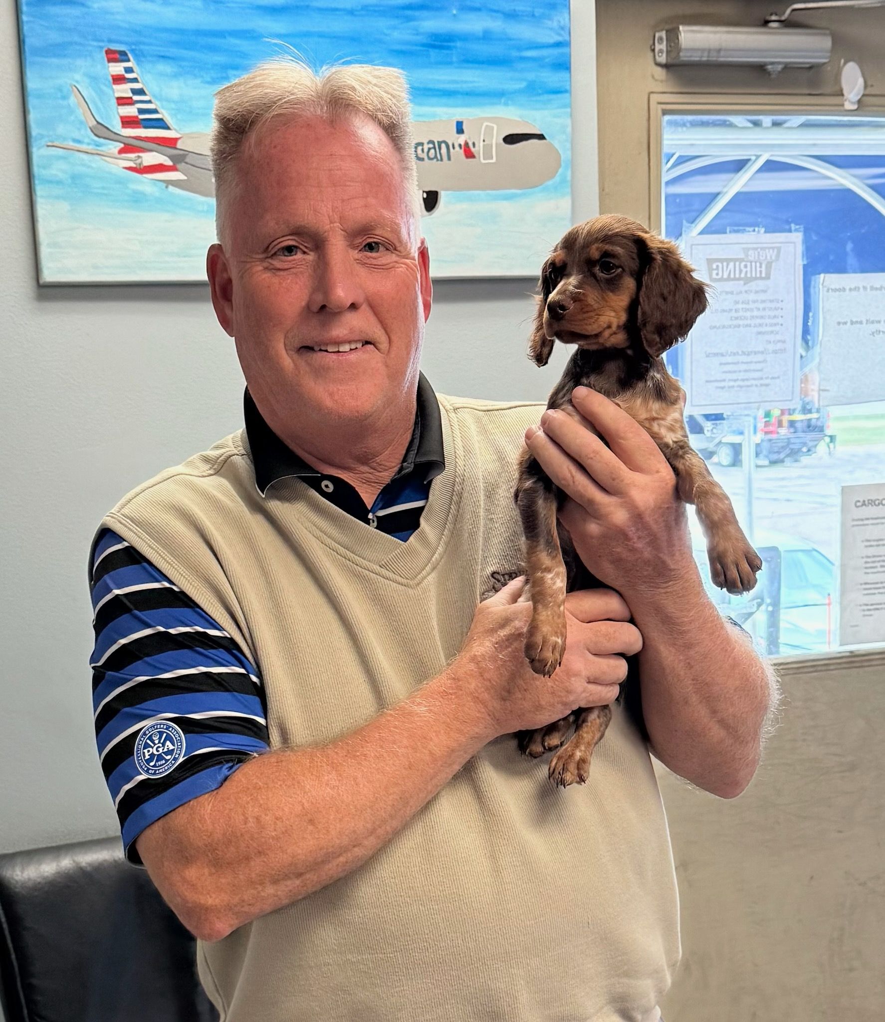 Man holding a small brown dog; painting of airplane in background.