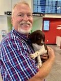 Man in plaid shirt holding a brown and white puppy; airport terminal background.