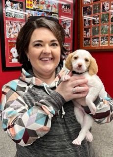 Woman holding a white and tan puppy, smiling. Red background with framed pictures.