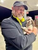 Man in a cap holds a small puppy, smiling. They are outside near a gas station at dusk.