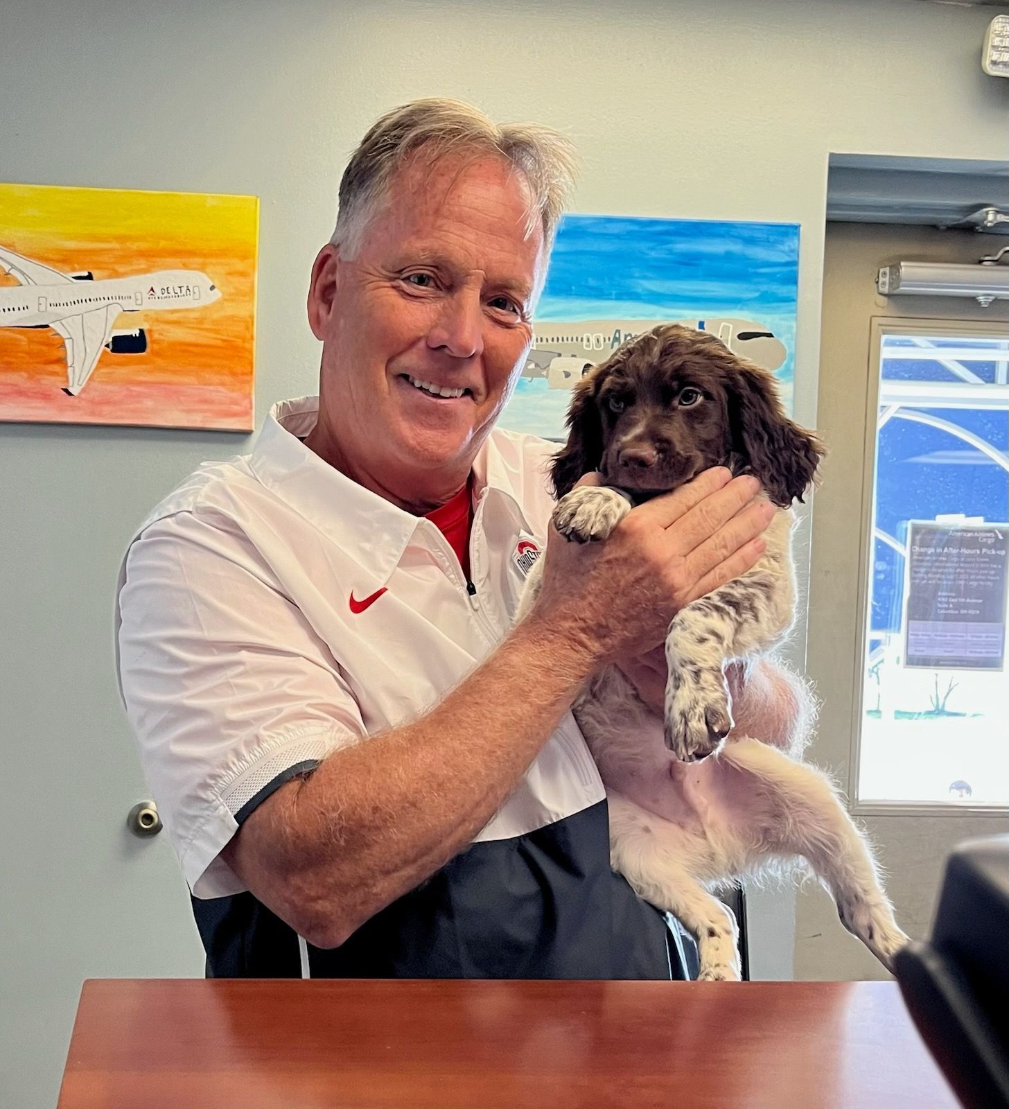 Man holding a small brown and white puppy, smiling. Artwork on the wall in the background.