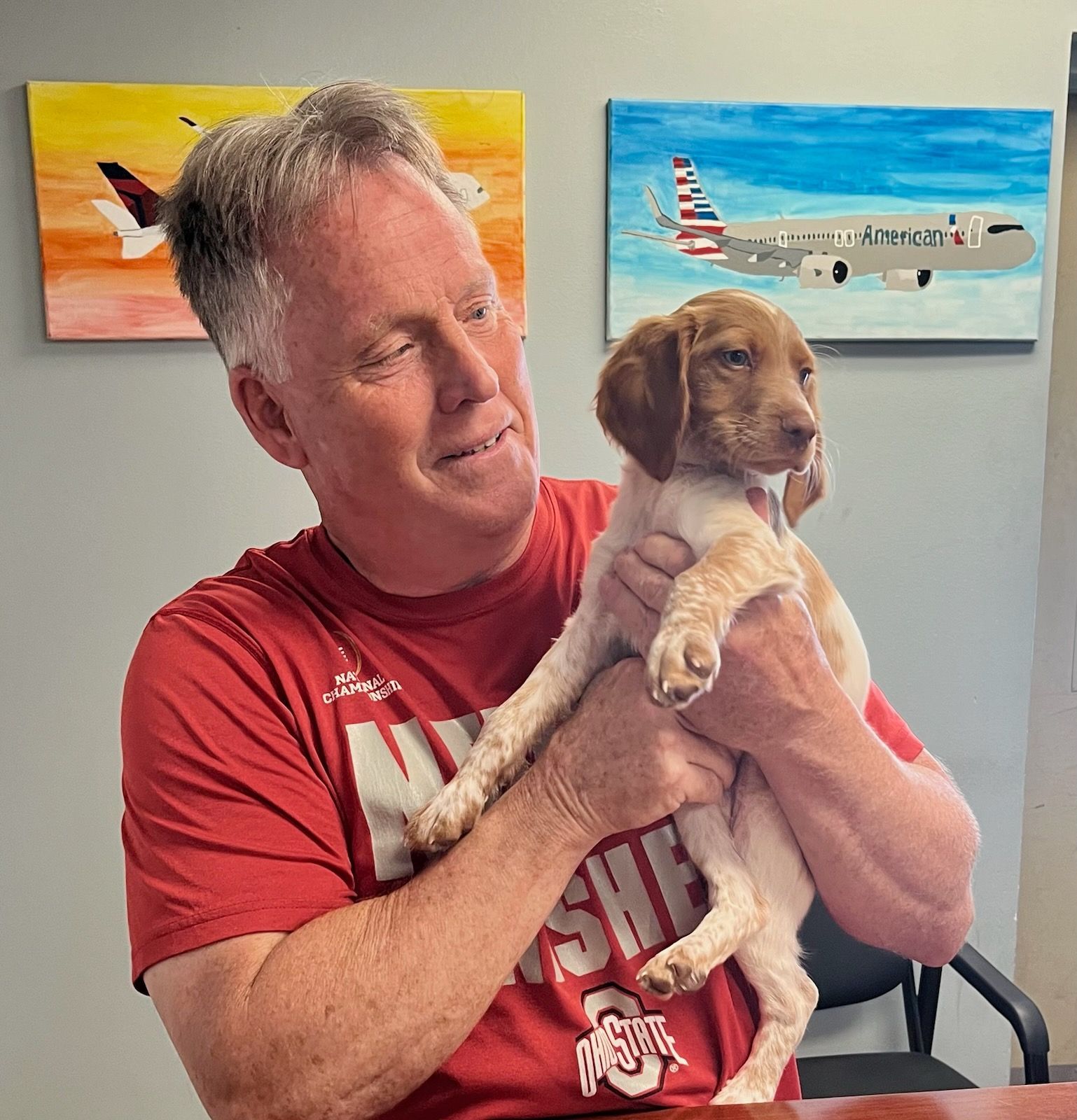 Man in red shirt holding a tan and white puppy, smiling. Paintings of airplanes on the wall.