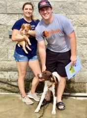 Couple with two dogs; one held, one on a leash. Outdoors, near a brick wall.
