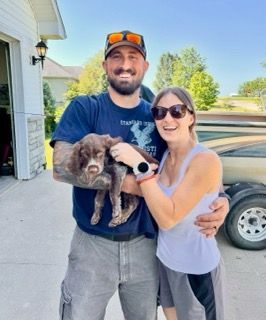 Man and woman smiling, holding a puppy outside by a garage.
