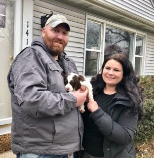 Couple holding a small brown and white puppy outside a house.