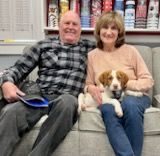 Couple and puppy on a couch. Man in plaid shirt, woman in pink, puppy in lap.