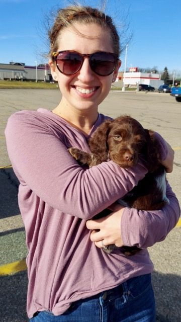 Woman in sunglasses hugs a brown puppy in an outdoor parking lot, smiling.