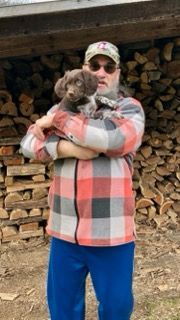 Man in plaid shirt and blue pants holding a puppy in front of a woodpile.