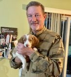 Man in camouflage shirt holding a beagle puppy; standing indoors.