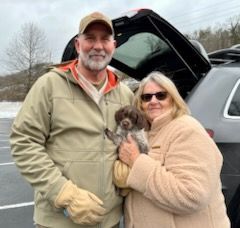 Couple with puppy near open car. Man wears cap, tan jacket. Woman wears coat, sunglasses. Puppy is brown and white.