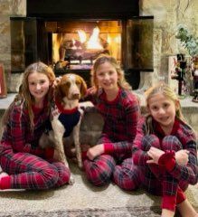 Three girls and a dog in matching red plaid pajamas sit in front of a fireplace.