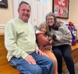Family sitting on a bench, holding a puppy; man in green sweater, girl in orange, woman in patterned coat.