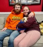Couple on couch cuddles puppy in blue jacket. Man in orange hoodie, woman in maroon sweatshirt. Red wall in background.