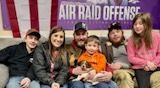 Family, a puppy, and friends seated together with a purple banner and an American flag in the background.