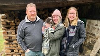 Family poses with a small dog in front of firewood. One person holds the puppy, smiling.