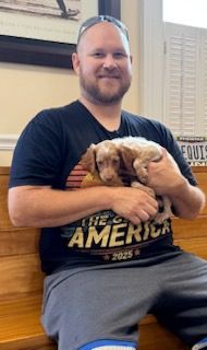 Man in black shirt holding a puppy. Both are inside, smiling.
