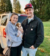 A couple holding a puppy outdoors. The woman is wearing a hoodie, the man a jacket and hat.