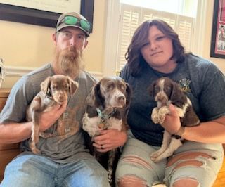 A man and a woman holding puppies indoors. The dogs are brown and white. They smile, posing.