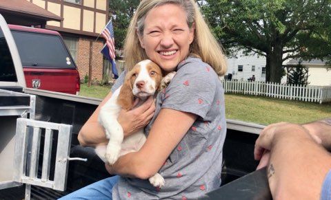 Woman in a pickup truck smiles and hugs a puppy with white and tan fur.