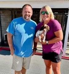 Man and woman smile, holding a small puppy outdoors near kennels.