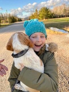 A child wearing a blue hat and green jacket hugs a small, spotted puppy with a big smile.