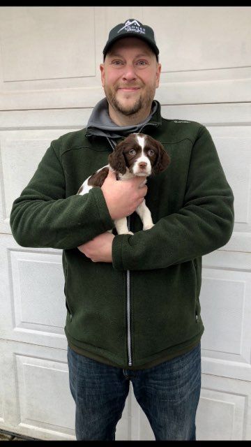 Man in green jacket and cap holding a brown and white puppy. Garage door in background.