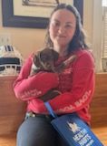 Woman in red sweater holds a small brown puppy on her lap, with a blue bag, indoors.