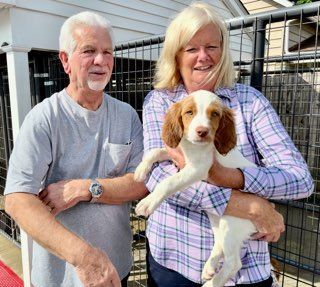 A couple holds a puppy in front of a fenced enclosure. The dog is white with brown ears.
