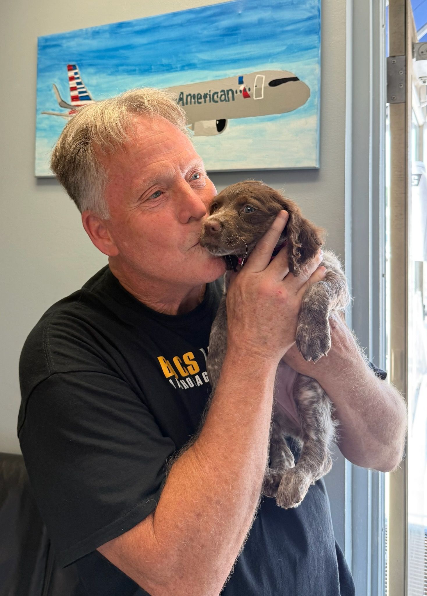 Man kissing a small brown and white dog. Painting of an airplane in the background.