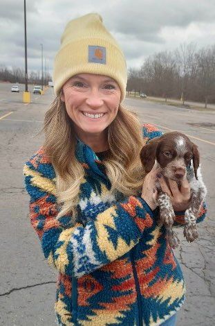 Woman in colorful jacket and yellow beanie holding a brown and white puppy in a parking lot.