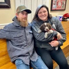 A man and woman sit, holding a puppy. The woman smiles, the man wears a hat. Inside.