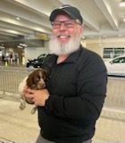 Man with a white beard holds a small brown and white puppy. Smiling in a parking garage.