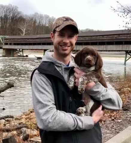 Man holding a brown and white puppy near a covered bridge over a river.