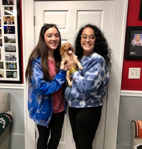 Two women, smiling, hold a small tan puppy in front of a white door. They wear tie-dye shirts.
