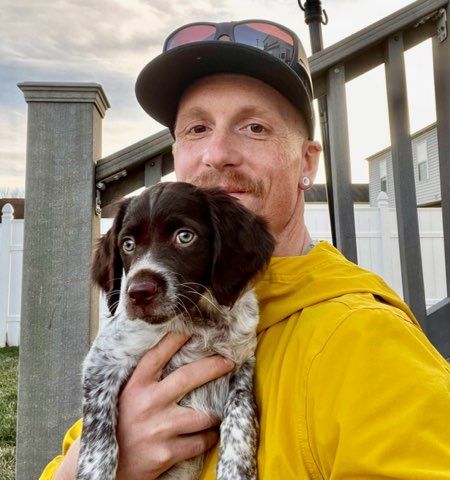 Man in yellow jacket holding a brown and white dog; outdoors, sunny.