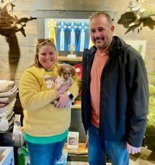 A woman and man smiling, holding a puppy in a shop. The woman wears yellow. The man wears a black jacket.