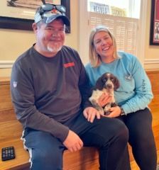 Couple sitting, holding puppy. Man in gray shirt and cap smiles, woman in blue shirt smiles. Wooden steps.