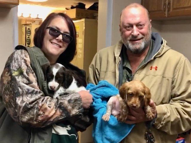 A couple holding two puppies inside a building. Woman in camo and man in tan coat smile.