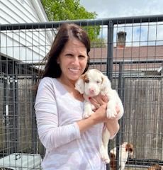 Woman holding a small white puppy with brown markings, smiling outside near a fenced enclosure.