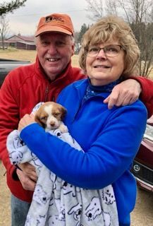 Couple holding a puppy wrapped in a blanket outdoors. Man smiles in red, woman smiles in blue, car visible.