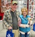 Man and woman smiling, holding small dog and blue tote bag; indoor setting.
