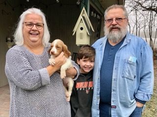 A smiling woman holds a puppy, a boy smiles beside her, and a man with a beard stands with them outdoors.